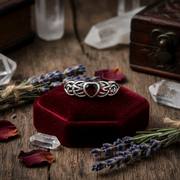 Silver ring with a heart-shaped gemstone on a red velvet squrae surrounded by crysals and herbs.