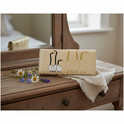 A cream-colored leather clutch wallet featuring a goose illustration, styled on a rustic wooden chest with a white fringed cloth and small daisies.