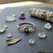Silver ring with a green gemstone on a piece of slate with sage, herbs and crystals displayed around it.