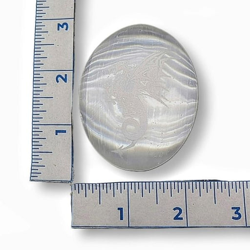 Selenite palm stone  with a etched dragon next to two rulers for size comparison on a white background