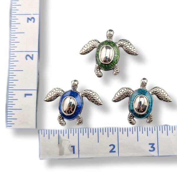 Three sea turtle jewelry charms with silver-toned metal bodies and enamel accents in green, blue, and turquoise, displayed on a white background with ruler measurements for scale.