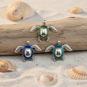 Three sea turtle jewelry pendants with silver-toned metal and colored enamel accents are arranged on a piece of driftwood over a sandy beach setting with seashells.
