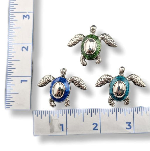 Three sea turtle jewelry charms with silver-toned metal bodies and enamel accents in green, blue, and turquoise, displayed on a white background with ruler measurements for scale.