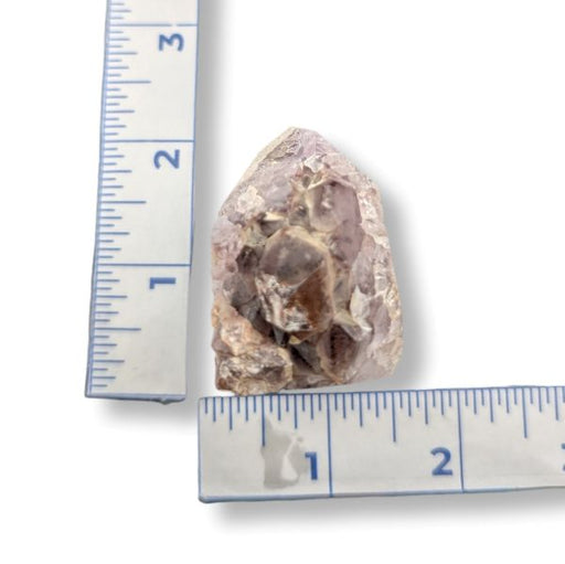 A rough, raw gemstone or mineral specimen with a rough, crystalline texture, shown next to two rulers (a centimeter ruler on the left and a metric ruler on the bottom) to indicate its size.