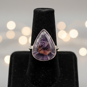 Teardrop-shaped amethyst ring on a black stand with blurred lights in the background