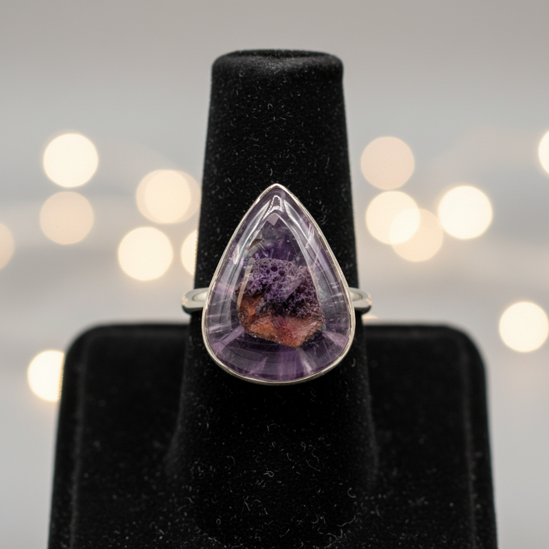Teardrop-shaped amethyst ring on a black stand with blurred lights in the background