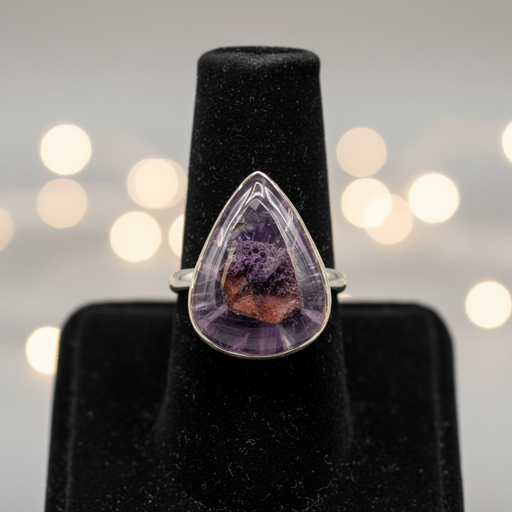 Teardrop-shaped amethyst ring on a black stand with blurred lights in the background