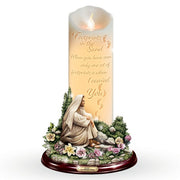 A devotional pillar candle with a printed image of the Virgin Mary kneeling in a garden, surrounded by flowers and greenery, with the text 'Footprints in the Sand' and 'When you have seen only one set of footprints is when I carried You' on the front, set against a plain white background.