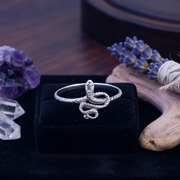 Silver snake-shaped ring on a black box with decorative elements like lavender and crystals.