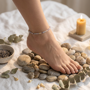 A close-up of a bare foot resting on smooth stones, wearing a delicate beaded ankle bracelet with a small gold charm. The scene is styled with eucalyptus leaves, a lit candle, and a small bowl of dark seeds on a white fabric background.