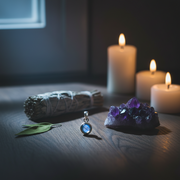Amethyst crystal, silver pendant, sage, and candles on a dark surface