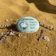 Decorative stone with Gemini zodiac information on a sandy background with lavender and crystals.
