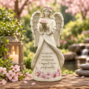 An angel memorial statue stands on a wooden surface in a softly blurred outdoor garden setting. The cream-colored angel figure has wings, a floral crown, and holds a small pink flower bouquet.