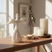 A white angel figurine stands on a wooden table in a softly lit room, styled with white pillar candles, a small tea light, dried flowers, and scattered petals.