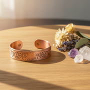 Copper bracelet with intricate designs on a wooden surface with flowers and crystals.