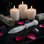 A white selenite spiritual knife with a carved handle rests on a black cloth surrounded by pink rose petals, purple stones, sage smudge bundles, and three lit white candles.