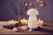 A selenite mushroom-shaped decorative object stands on a wooden surface, surrounded by pink and purple crystals, lit candles, a reed diffuser with incense sticks, and a smudge bundle against a soft purple bokeh background.