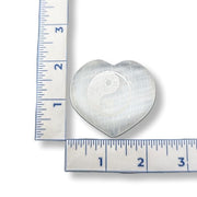 Heart-shaped selenite with a yin yang symbol next to a ruler for scale on a white background
