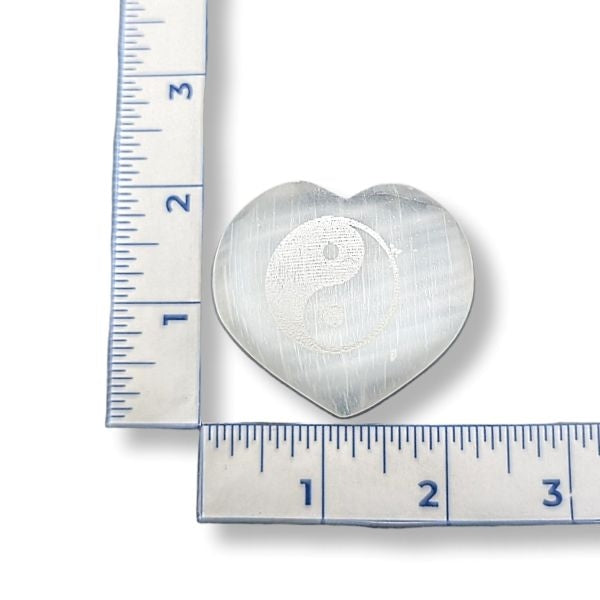 Heart-shaped selenite with a yin yang symbol next to a ruler for scale on a white background