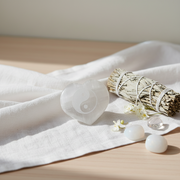 A heart-shaped selenite with a yin-yang symbol inside, styled on a white linen cloth next to a dried sage smudge bundle and small white stones on a light wood surface.