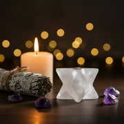 A Selenite Merkaba sits on a dark wooden surface next to a white candle, a sage smudge bundle, and purple amethyst stones, with warm bokeh lights in the background.
