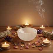 A selenite bowl sits on a wooden table surrounded by lit tealight candles, smudge bundles, dried petals, crystals, and incense sticks in a warm, atmospheric arrangement.