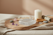 A square selenite dish sits on a round wooden tray, surrounded by crystals, a white candle, a sage bundle, and smooth stones on draped beige fabric.
