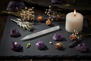 A white taper candle burns on a dark slate tray surrounded by purple amethyst crystals, dried roses, baby's breath, lavender, and a white selenite pencil wand.