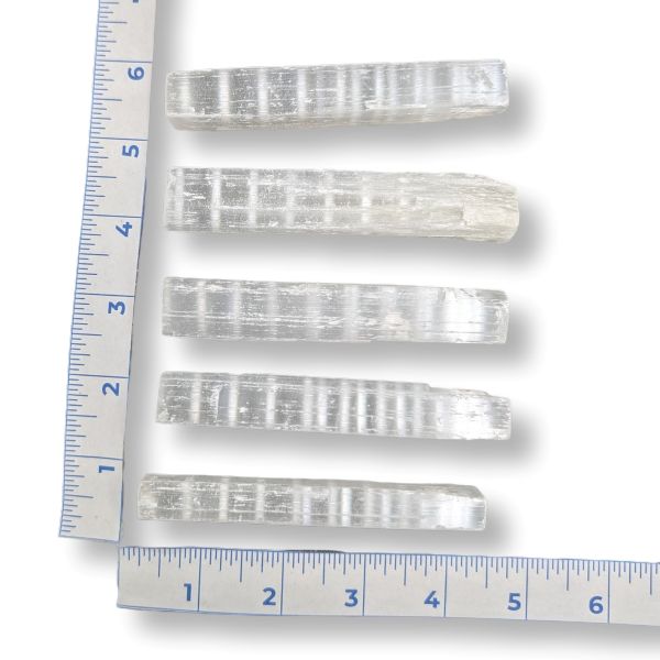 Five clear, ribbed selenite sticks arranged vertically next to a blue ruler for scale, on a white background.