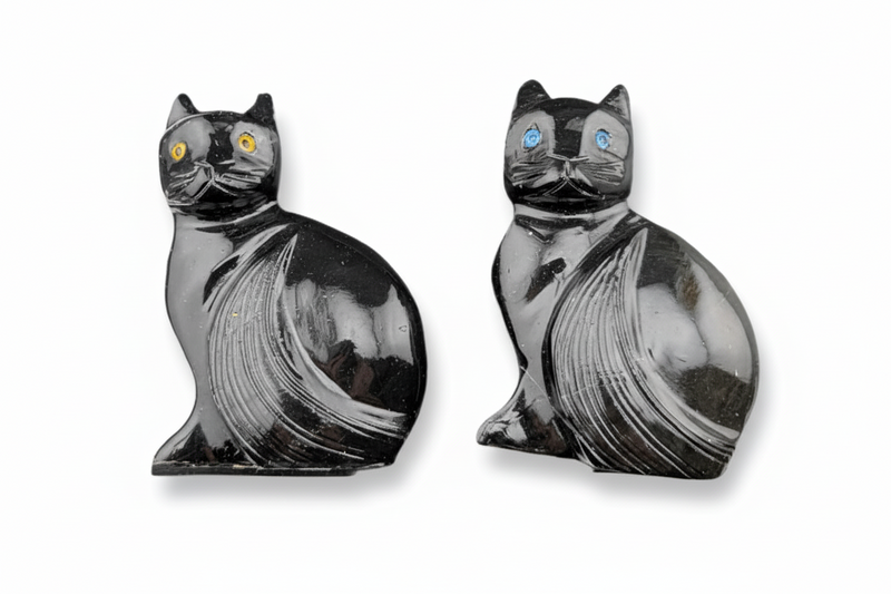 Two polished black cat-shaped carving s with carved fur details and one featuring yellow-gold and the other blue inlaid eyes, displayed side by side on a white background.