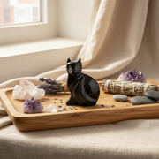 A glossy black cat figurine sits on a wooden tray surrounded by crystals, dried lavender, a sage bundle, and smooth stones, set against a soft beige curtain and window background.