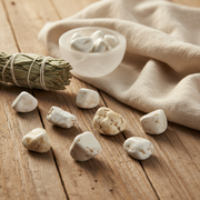 White stones on a wooden surface with a bundle of sage and a cloth in the background.