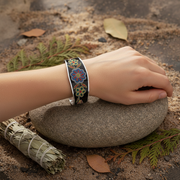 A silver-toned cuff bracelet with a colorful floral inlay is worn on a person's wrist, resting on a smooth gray stone outdoors with sand, leaves, and a bundle of dried leaves nearby.