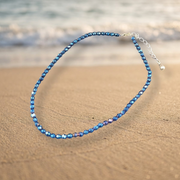 A beaded anklet with faceted blue beads and a silver clasp, photographed on a sandy beach with the ocean blurred in the background.