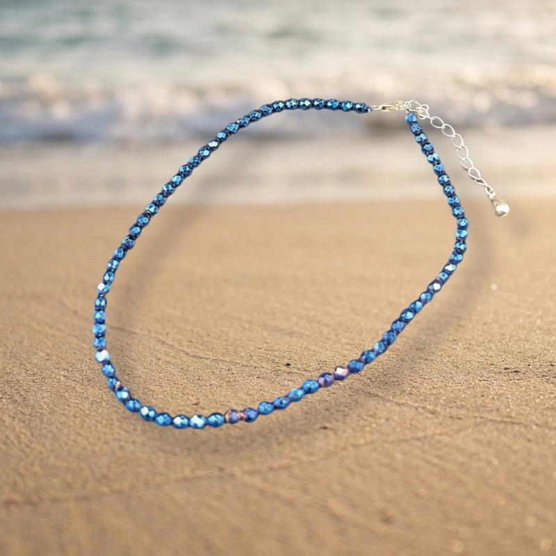 A beaded anklet with faceted blue beads and a silver clasp, photographed on a sandy beach with the ocean blurred in the background.
