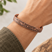 A close-up of a person's wrist wearing a copper-toned engraved bracelet with dark recessed detailing and small dark metal rivets, against a softly blurred light background.