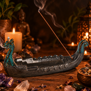 A decorative boat-shaped incense holder with a dragon head, shown on a wooden surface with a lit incense stick, candles, crystals, and warm ambient lighting in a styled indoor setting.