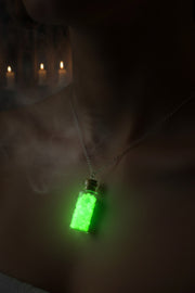 A glowing green potion bottle pendant necklace worn on a person's neck, with a dark background and blurred candles in the distance.