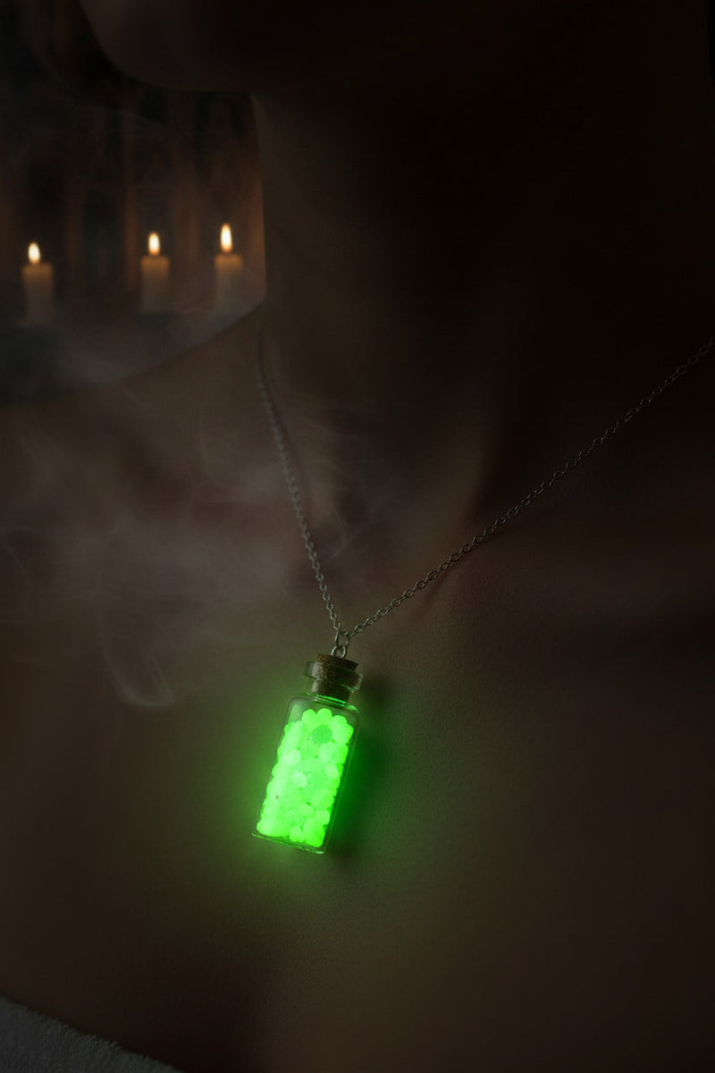 A glowing green potion bottle pendant necklace worn on a person's neck, with a dark background and blurred candles in the distance.
