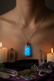 A silver chain necklace with a small glowing blue vial pendant displayed on a model's chest, surrounded by candles, crystal points, dried herbs, and pentagram charms on a purple cloth.