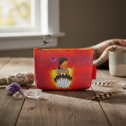 A small zip pouch with a colorful spiritual illustration of a woman holding feathers, set on a rustic wooden table with crystals, a smudge bundle, and a mug in the background.