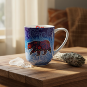 A ceramic mug with a blue and purple bear design sits on a wooden table next to a sage smudge and crystals, with a warm, softly blurred background.