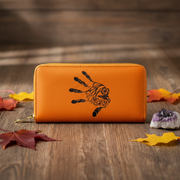 An orange pebbled leather zip-around wallet with a black handprint design, displayed on a rustic wooden surface with autumn leaves and crystals.