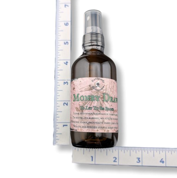 An amber glass spray bottle labeled 'Money Draw' shown next to a blue measuring tape ruler on a white background.
