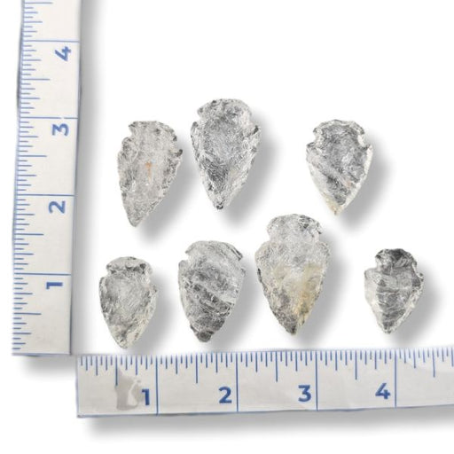 Six small pointed stone arrowheads arranged on a white background with a measuring ruler along the left and bottom edges for scale.