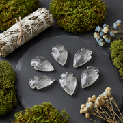 Five clear crystal leaf-shaped arrowheads arranged on a dark slate board, surrounded by green moss, dried flowers, and a white sage smudge bundle.