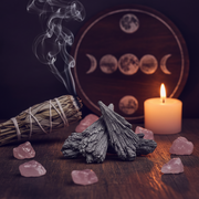A styled tabletop scene with a bundle of dried smudge sticks smoking on the left, a small stack of black kyanite, scattered pink stones, and a lit white candle on the right. A round wooden board with moon phases sits blurred in the background against a dark setting.