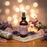 An amber glass spray bottle labeled 'Be Gone' stands on a patterned cloth on a wooden surface, surrounded by crystals, a smudge bundle, candles, and dried flowers against a warm bokeh background.