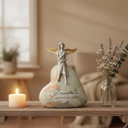 A heart-shaped statue with a silver angel figurine sitting on top, printed with the words 'Grandma ALWAYS MY ANGEL.' The urn is styled on a wooden shelf next to a lit candle and a glass vase with dried flowers in a warm, softly blurred room.