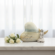 Heart-shaped memorial stone with a painted ocean scene and the words 'In Loving Memory' on a white marble surface, styled with white roses and a small kneeling angel figurine.